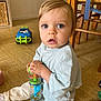 toddler, child, toy, floor, blue_eyes, curious, indoor, clothing, pants, shirt, plastic_toy, colorful, furniture, chair, wooden, playtime, expression, hands, face, home