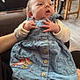 baby, infant, child, denim_dress, blue_dress, floral_pattern, bird_embroidery, hand, curious, indoor, flooring, wooden_chair, seated, cute, portrait, clothing, person, skin, face, newborn