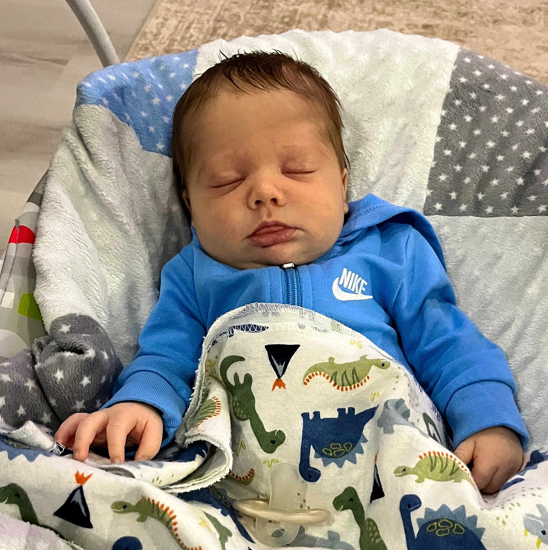 Luka is registered to the contest to win money with this photo: baby, sleeping, blanket, dinosaur_pattern, blue_hoodie, nike_logo, infant, child, cushion, baby_seat, hand, pacifier, quilt, star_pattern, indoor, cozy, resting, cute, young_child, soft_fabric