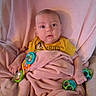 baby, blanket, toy, yellow_shirt, cushion, chair, infant, indoor, face, person, child, comfort, soft, cute, expression, holding, colorful, pink, seated, curious