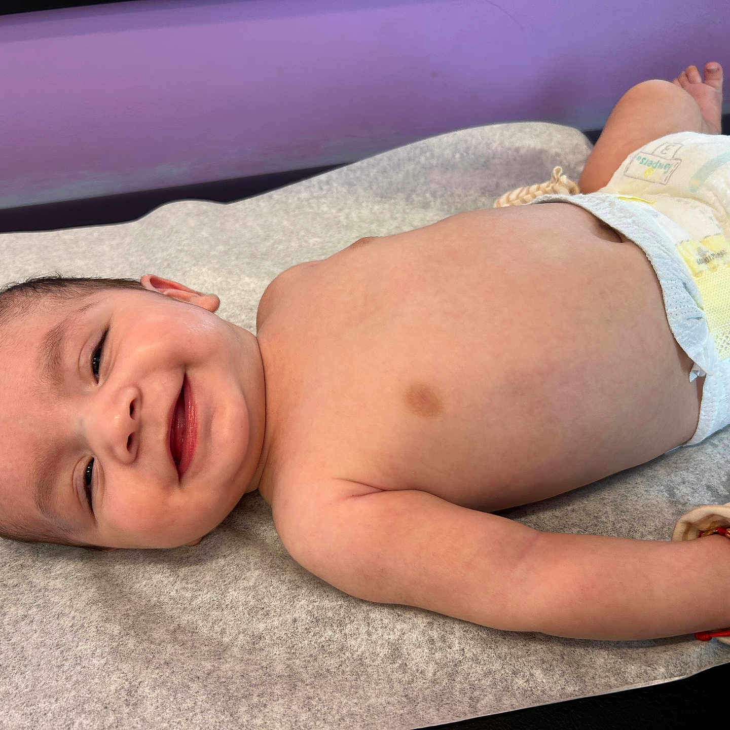 Luka joined the competition — help win amazing prizes! baby, infant, child, smiling, lying_down, diaper, changing_mat, portrait, face, eyes, hair, skin, bracelet, indoor, close_up, happy, relaxed, nursery, purple_background, photography