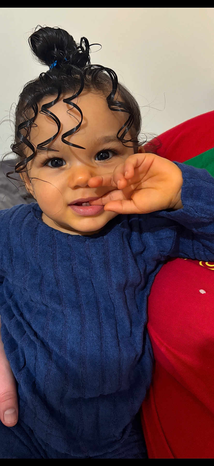 Maëna a rejoint le concours — aidez-le/la à gagner de superbes lots ! child, toddler, face, eyes, curly_hair, hair_bun, hand, finger, blue_sweater, smile, eyelashes, cheeks, lips, indoors, couch, red_cushion, portrait, close_up, skin, cute