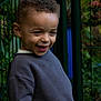 child, smiling, curly_hair, sweater, toy, green_fence, outdoor, nature, plants, happy, portrait, young, playful, person, casual, daylight, cute, expression, boy, leisure