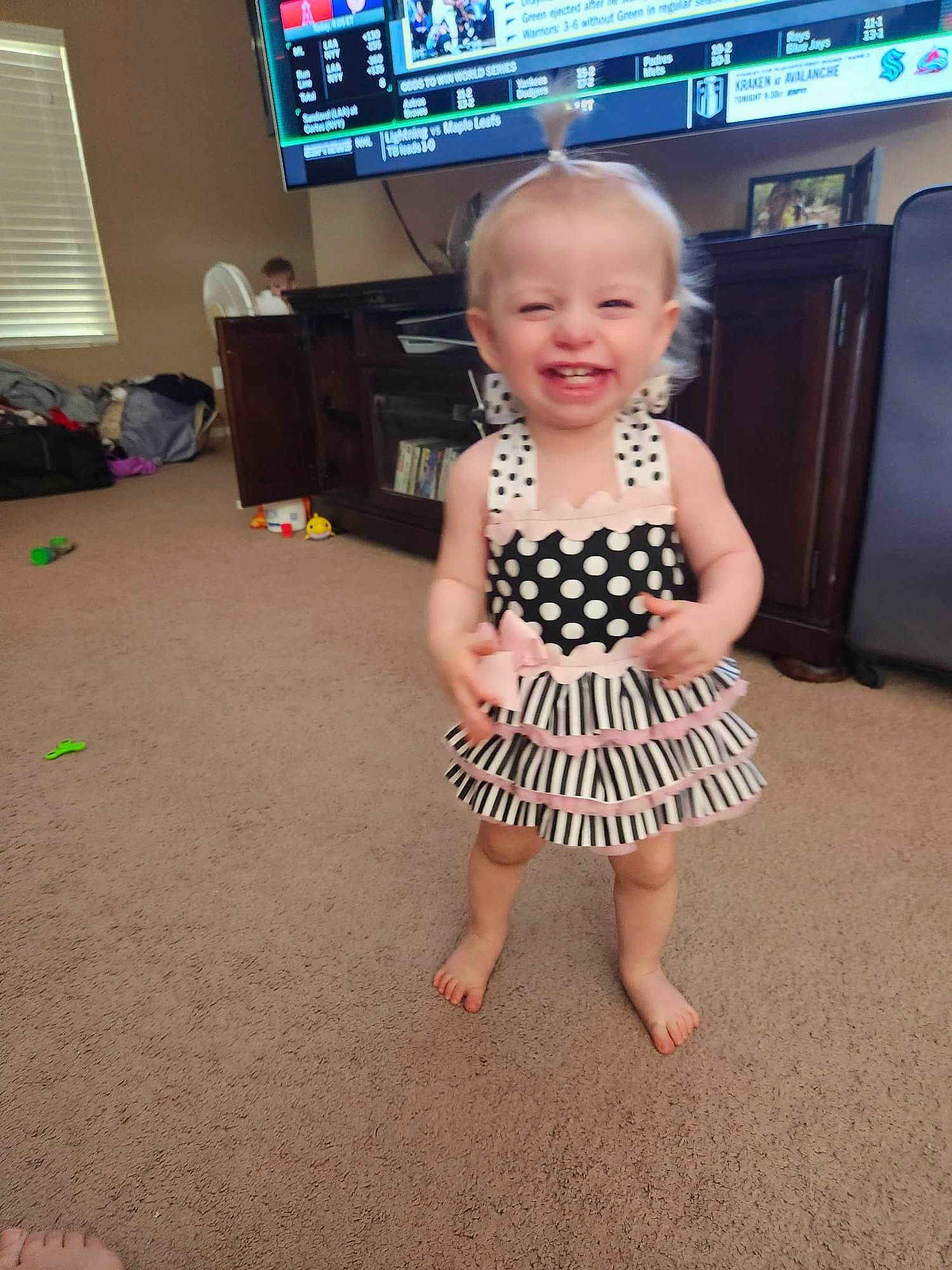 Gracelynn is registered to the contest to win money with this photo: baby_toddler_clothing, child, day_dress, finger, floor, flooring, foot, fun, happy, hardwood, home_appliance, joint, joy, one_piece_garment, person, smile, standing, television, television_set, thumb