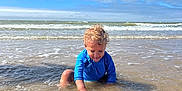 Lowen participe au concours pour gagner de l'argent avec cette photo : child, toddler, ocean, water, beach, waves, blue_sky, clouds, curly_hair, blue_clothing, play, sitting, sunlight, sand, shallow_water, happy, tongue_out, summer, outdoor, nature