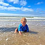 child, toddler, ocean, water, beach, waves, blue_sky, clouds, curly_hair, blue_clothing, play, sitting, sunlight, sand, shallow_water, happy, tongue_out, summer, outdoor, nature