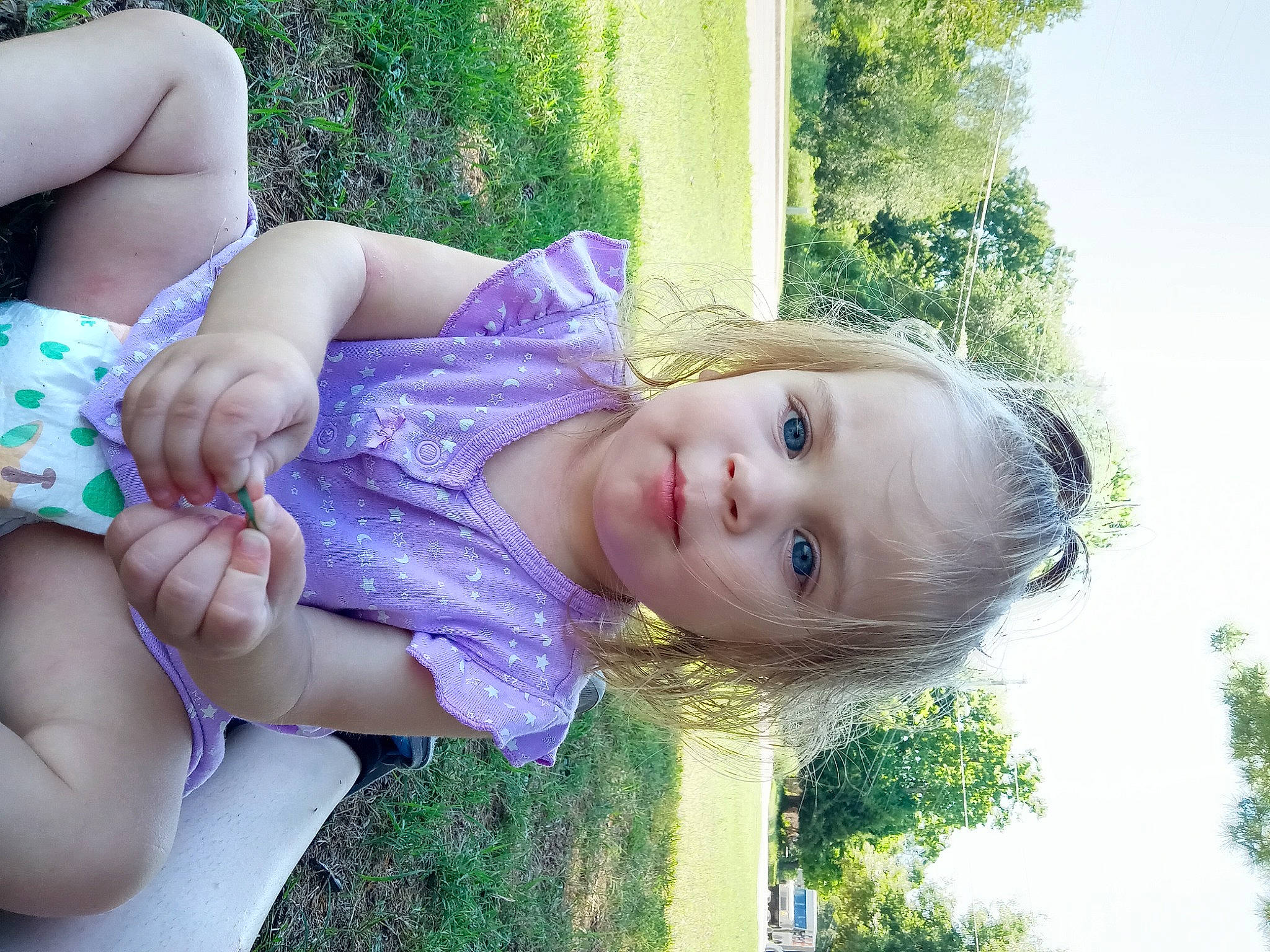 Mallory is registered to the contest to win money with this photo: baby_toddler_clothing, child, facial_expression, fun, grass, green, happy, human_body, iris, leisure, lip, people_in_nature, person, plant, sitting, skin, sky, smile, toddler, tree