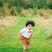 Zayan is registered to the contest to win money with this photo: toddler, child, curly_hair, white_shirt, brown_shorts, suspenders, bow_tie, grass, greenery, outdoor, nature, pathway, trees, grass_field, portrait, cute, young_child, standing, curious, daylight