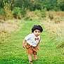 toddler, child, curly_hair, white_shirt, brown_shorts, suspenders, bow_tie, grass, greenery, outdoor, nature, pathway, trees, grass_field, portrait, cute, young_child, standing, curious, daylight