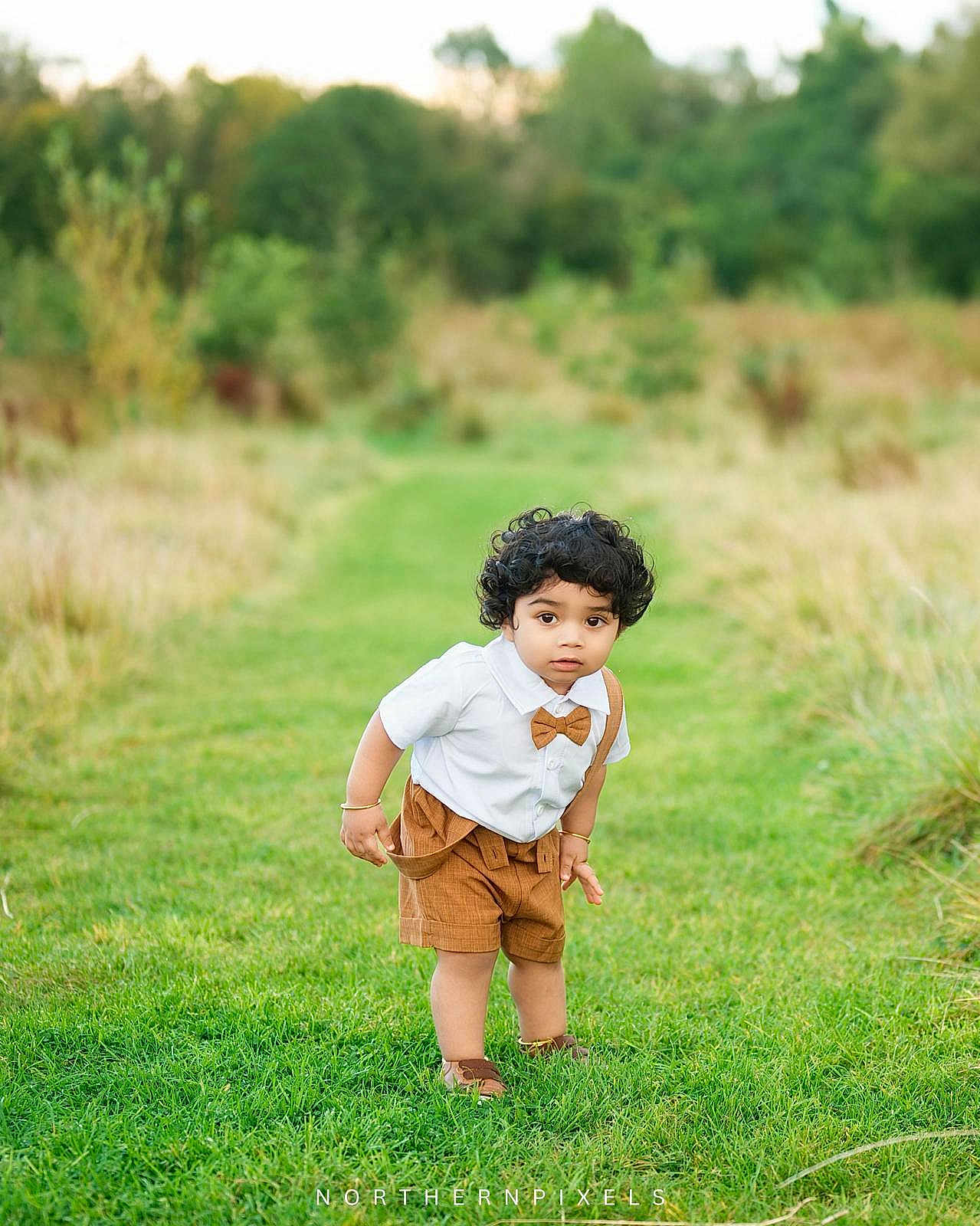 Zayan is registered to the contest to win money with this photo: toddler, child, curly_hair, white_shirt, brown_shorts, suspenders, bow_tie, grass, greenery, outdoor, nature, pathway, trees, grass_field, portrait, cute, young_child, standing, curious, daylight