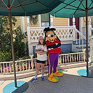 Julie a rejoint le concours — aidez-le/la à gagner de superbes lots ! child, person, costume, cartoon_character, umbrella, outdoor, smile, standing, flowerbed, house, railing, sidewalk, hat, shorts, tshirt, shoes, character, theme_park, daytime, fun