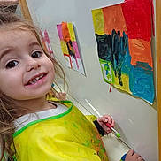 Mia a rejoint le concours — aidez-le/la à gagner de superbes lots ! child, girl, painting, art, smile, yellow_smock, brush, colorful, wall, happy, creative, indoor, young_artist, portrait, expression, person, drawing, paint, hands, artwork
