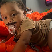 Robert joined the competition — help win amazing prizes! baby, background, child, clothing, curious, cute, expression, face, indoor, infant, lying_down, mustache, orange_blanket, pacifier, person, reaching, room, soft_lighting, toddler, toy