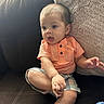 Nathan is registered to the contest to win money with this photo: baby, child, infant, couch, sofa, pillow, orange_shirt, plaid_shorts, sitting, indoors, curious, expression, cute, young_child, furniture, home, casual_clothing, skin, hand, foot