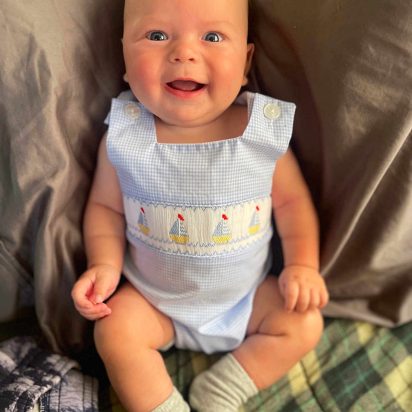 Maddox is registered to the contest to win money with this photo: baby, blanket, blue_eyes, child, closeup, comfortable, cute_outfit, happy, homedecor, indoor, infant, pillow, plaid_blanket, portrait, sailboat_pattern, sitting, skin, smiling, socks, soft_lighting