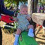 Maddox is registered to the contest to win money with this photo: baby, child, motorbike, green, outdoor, person, glove, hand, seat, socks, overalls, helmet, dirt, sunlight, tree, fence, people, daytime, serious, blurred_background