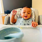 Leo is registered to the contest to win money with this photo: baby, bib, bowl, child, cute, early_childhood, feeding_time, food, happy, high_chair, home, indoor, infant, kitchen, messy_face, person, portrait, smiling, spoon, waving_hand