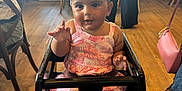 Natalia is registered to the contest to win money with this photo: baby, child, high_chair, bow, pink_dress, indoor, wooden_floor, restaurant, people, woman, man, furniture, curious, cute, person, seated, smiling, headband, toddlers, family