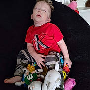 Caden is registered to the contest to win money with this photo: toddler, child, sleeping, red_shirt, camouflage_pants, stuffed_toys, plush, chair, indoors, relaxed, cute, nap, person, young_child, soft_toys, black_background, casual_clothing, resting, small_person, cozy