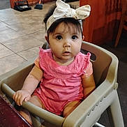 Juanita Quiroz is registered to the contest to win money with this photo: baby, child, high_chair, pink_clothing, headband, bow, indoor, floor_tiles, table, wooden_furniture, face, person, curious, seated, sock, cleaning_tools, broom, dustpan, expression, wide_eyes