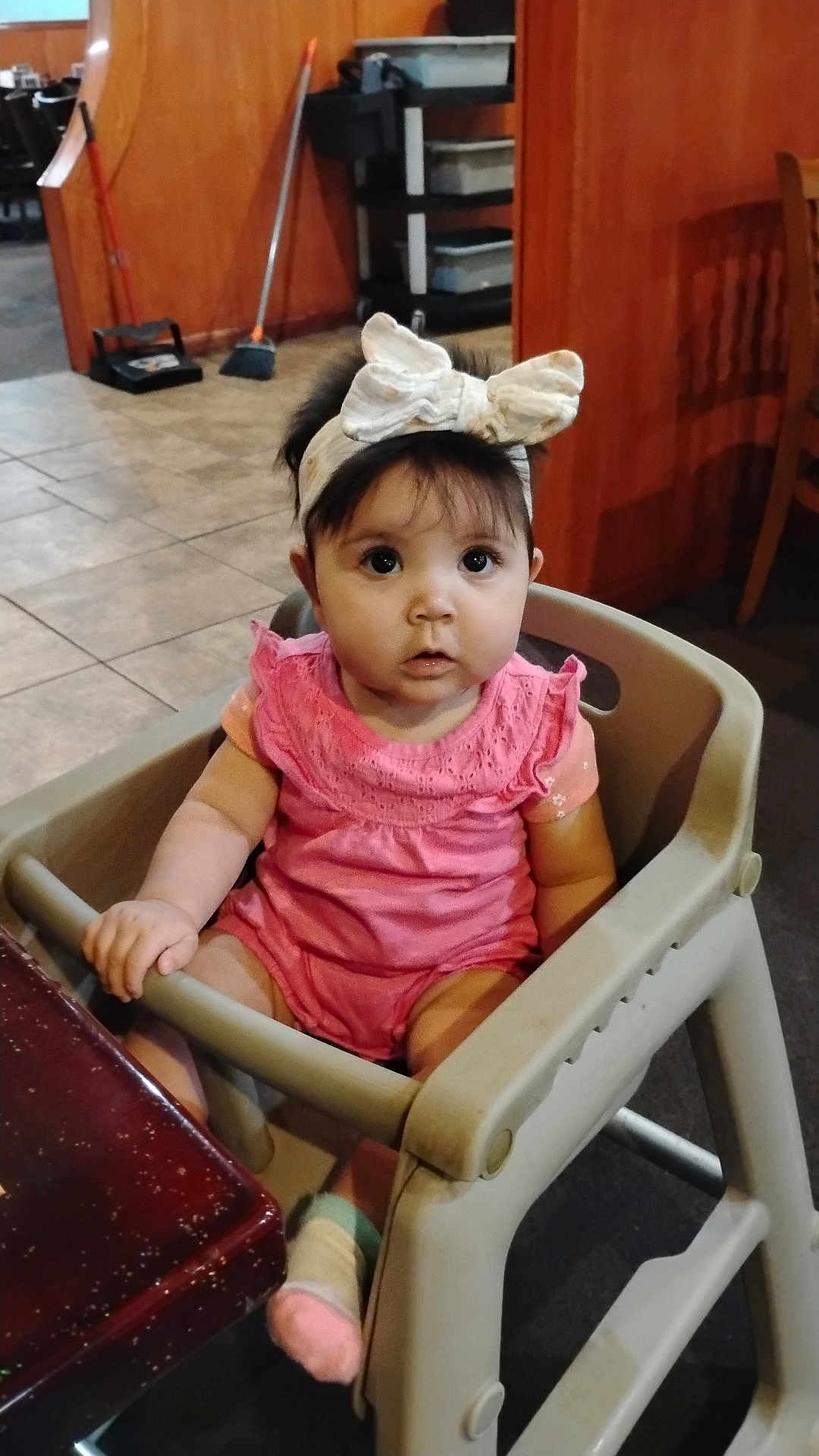 Juanita Quiroz is registered to the contest to win money with this photo: baby, child, high_chair, pink_clothing, headband, bow, indoor, floor_tiles, table, wooden_furniture, face, person, curious, seated, sock, cleaning_tools, broom, dustpan, expression, wide_eyes