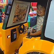 Zohan is registered to the contest to win money with this photo: child, boy, arcade, ride, yellow, smile, game, indoor, entertainment, machine, joy, happiness, play, seat, steering_wheel, screen, toy, fun, casual_clothing, footwear