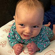 Rose is registered to the contest to win money with this photo: baby, infant, smiling, face, hands, clothing, floral_pattern, cushion, heart_pattern, lying_down, cute, child, person, indoors, skin, head, eyes, fingers, happy, toddler
