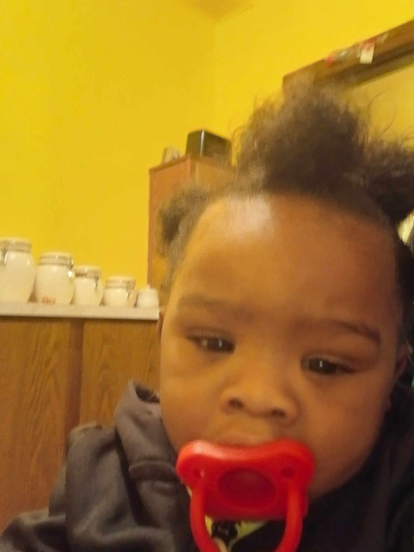Kayden is registered to the contest to win money with this photo: baby, pacifier, indoor, child, face, hair, cabinet, wall, yellow, clothing, furniture, room, head, portrait, infant, young_child, person, closeup, blur, expression