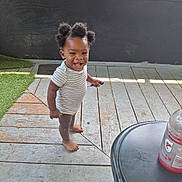 Kayden joined the competition — help win amazing prizes! toddler, child, barefoot, smiling, hair_puffs, striped_onesie, wooden_deck, outdoor, pen, table, beverage_bottle, window, green_turf, black_wall, sunlight, happy, person, standing, casual, daylight