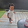toddler, child, barefoot, smiling, hair_puffs, striped_onesie, wooden_deck, outdoor, pen, table, beverage_bottle, window, green_turf, black_wall, sunlight, happy, person, standing, casual, daylight