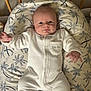 baby, infant, onesie, white_clothing, cushion, palm_tree_pattern, wooden_bed, blue_eyes, lying_down, cute, chubby_cheeks, hand, face, portrait, indoors, soft_lighting, baby_bed, sleepwear, newborn, child