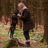 Raykon a rejoint le concours — aidez-le/la à gagner de superbes lots ! woman, dog, forest, tree_stump, moss, pine_cones, outdoor, nature, black_dog, glasses, coat, boots, interaction, pet, affection, trees, ground, standing, smile, leaves