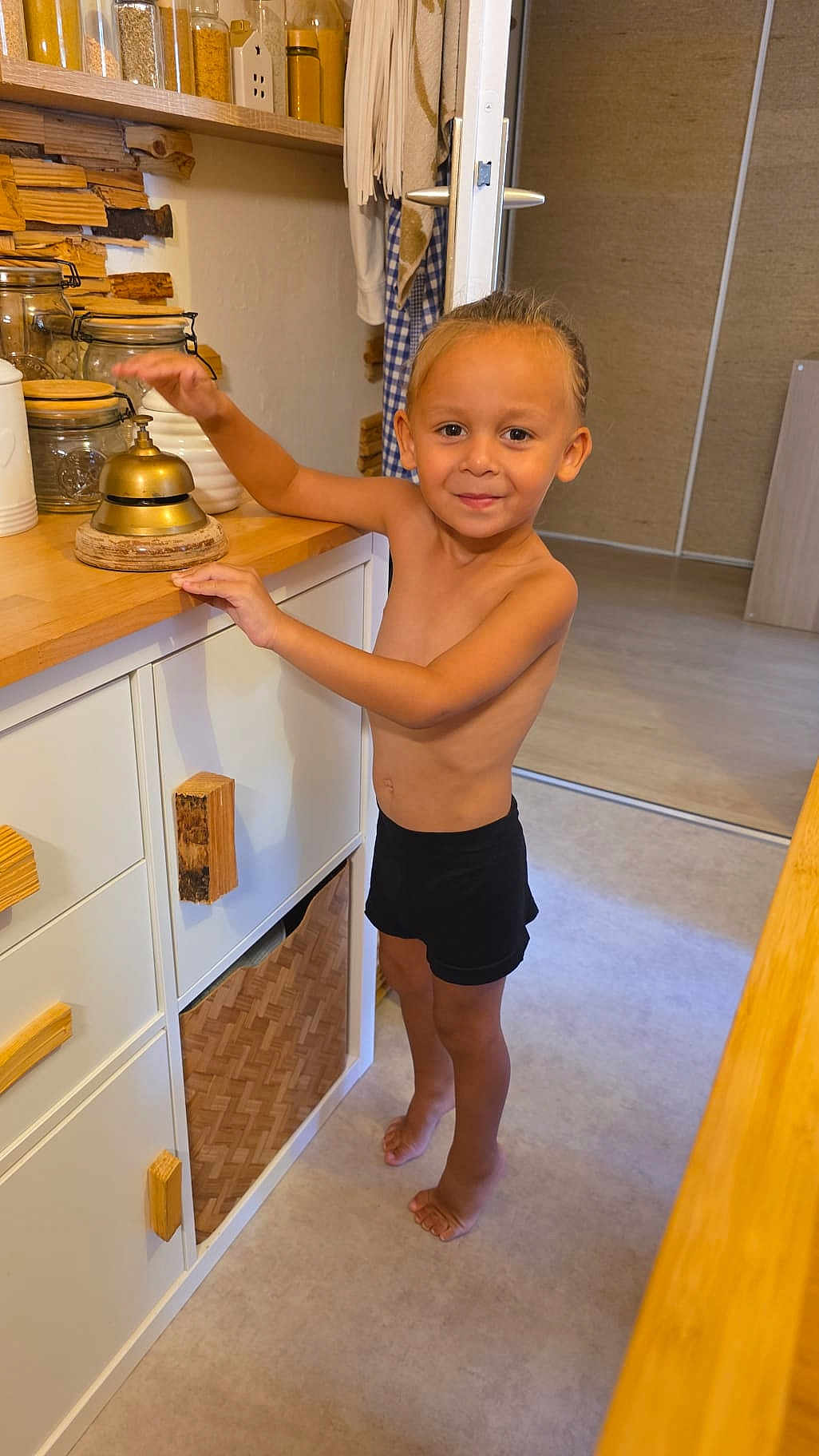 Waël a rejoint le concours — aidez-le/la à gagner de superbes lots ! toddler, child, barefoot, smiling, braid, kitchen, bell, cabinet, wood, countertop, jar, floor, home, interior, person, furniture, young_child, standing, cute, happy