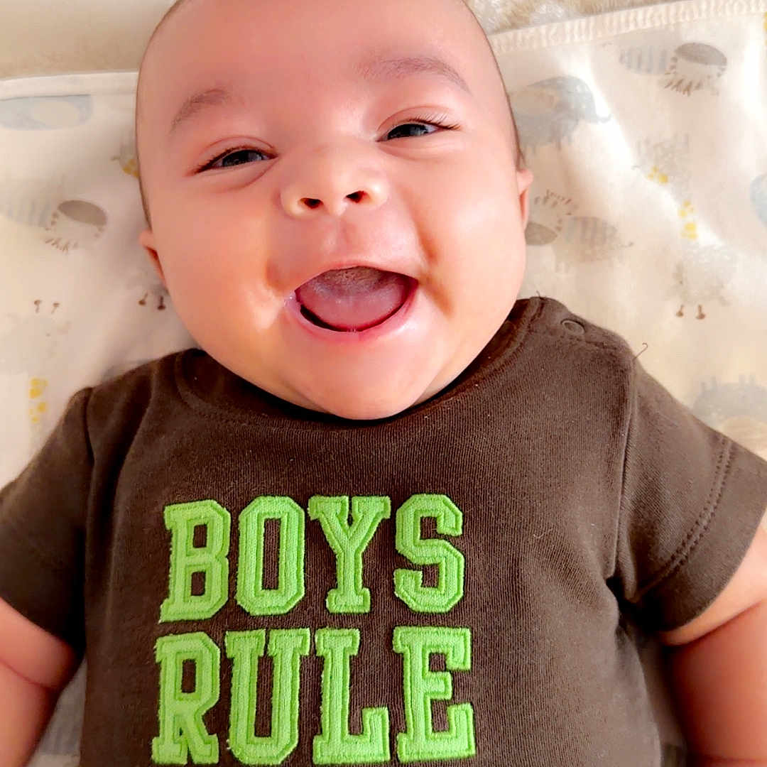 Zayden is registered to the contest to win money with this photo: baby, smiling, infant, onesie, clothing, face, happy, lying_down, child, cute, apparel, text_on_clothing, skin, human, portrait, toddler, baby_boy, closeup, indoor, blanket