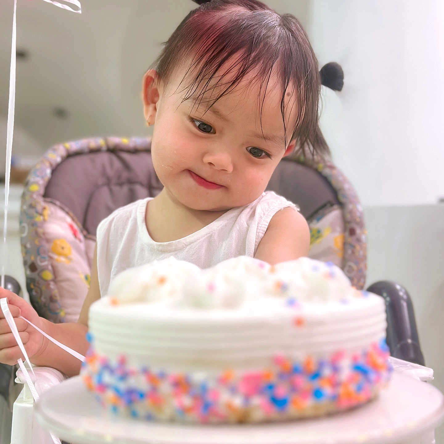 Janira Truong is registered to the contest to win money with this photo: birthday_cake, cake, celebration, child, curly_hair, expression, face, food, high_chair, holding, indoor, party, person, pigtails, plate, sprinkles, sweet, toddler, white_clothing, white_frosting
