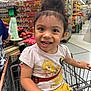 child, toddler, smiling, shopping_cart, grocery_store, clothing, hair_bun, yellow_shorts, princess_tshirt, face, person, happy, indoor, aisle, food, produce, retail, store, cute, seatbelt
