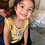 Jasmine is registered to the contest to win money with this photo: child, smiling, portrait, indoors, bed, pillow, blanket, happy, person, casual_clothing, yellow_shirt, black_shorts, cute, young, girl, hair, face, sitting, home, cozy