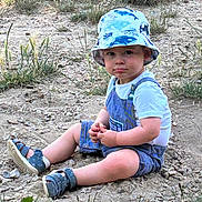 Elliot a rejoint le concours — aidez-le/la à gagner de superbes lots ! bucket_hat, casual_clothing, child, curious, daylight, dirt, exploration, grass, hands, hat, nature, outdoor, overalls, portrait, rocks, sandals, sitting, summer, toddler, young_child