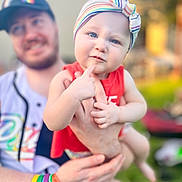 Dakoda joined the competition — help win amazing prizes! baby, child, adult, person, headband, rainbow, colorful, wristbands, red_clothing, outdoor, greenery, smiling, holding, portrait, face, hand, curious_expression, background_blur, sunlight, casual