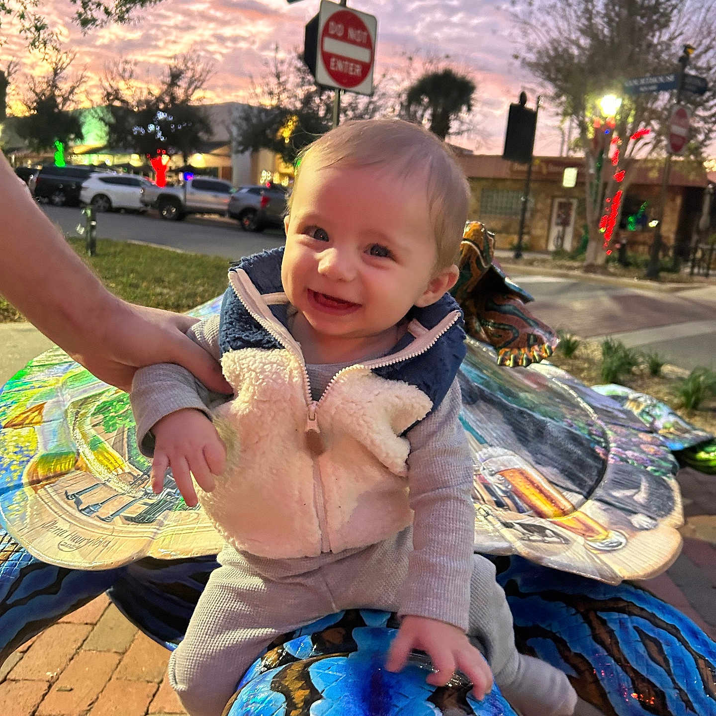 Rippley is registered to the contest to win money with this photo: baby, brick_pavement, cars, child, colorful, cute, dusk, hand, happy, lights, outdoor, person, portrait, sculpture, sign, smiling, street, trees, turtle, vest