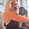 Monster is registered to the contest to win money with this photo: affection, arm, black_and_white_dog, blonde_hair, casual_clothing, chair, comfort, dog, glasses, home, hugging, indoor, love, natural_light, pet, portrait, relaxed, seated, window, woman