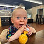 Kevin is registered to the contest to win money with this photo: baby, child, toy, high_chair, indoor, table, blue_eyes, colorful_clothing, wooden_table, chewing, happy, cute, wild_hair, restaurant, menu_board, chair, floor, lighting, face, person