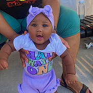 Zaierra is registered to the contest to win money with this photo: adult_hands, baby, bench, black_history, bottle, child, colorful_clothing, concrete_floor, happy, headband, outdoor, person, purple_outfit, sandals, shirt, smiling, standing, sunglasses, support, table