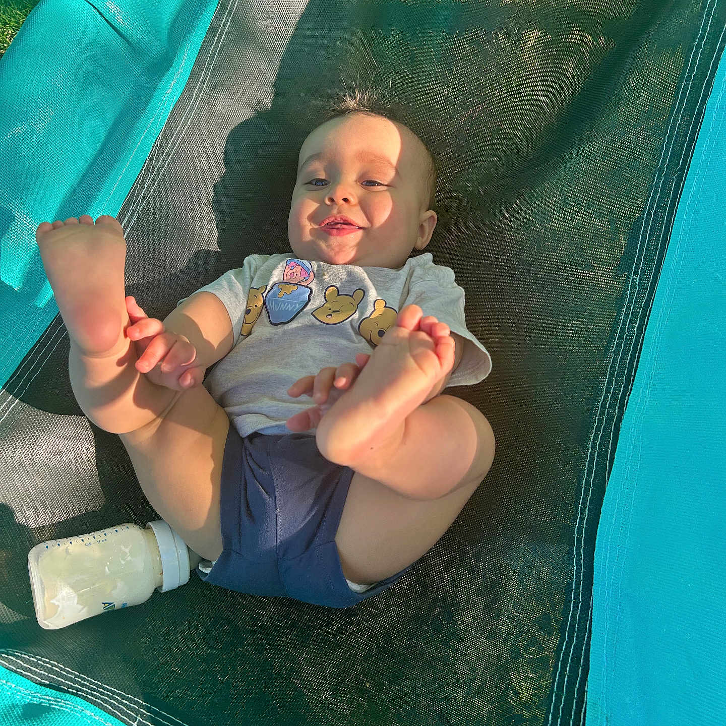 Domari is registered to the contest to win money with this photo: baby, child, smiling, feet, milk_bottle, outdoor, sunlight, mesh, teal, black, clothing, tshirt, shorts, grass, playful, infant, person, happy, relaxing, summer
