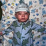 Omar is registered to the contest to win money with this photo: baby, infant, dinosaur_onesie, hat, blanket, toddler, cute, sleepwear, child, portrait, face, expression, patterned_clothing, soft_texture, lying_down, indoor, cozy, colorful, animal_print, newborn