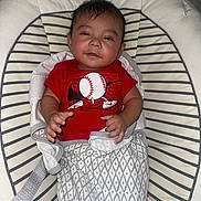 Omar joined the competition — help win amazing prizes! baby, infant, child, red_shirt, baseball_graphic, striped_cushion, baby_seat, blanket, patterned_blanket, indoor, smiling, person, clothing, cute, comfort, resting, face, hand, hair, wall