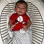 baby, infant, child, red_shirt, baseball_graphic, striped_cushion, baby_seat, blanket, patterned_blanket, indoor, smiling, person, clothing, cute, comfort, resting, face, hand, hair, wall