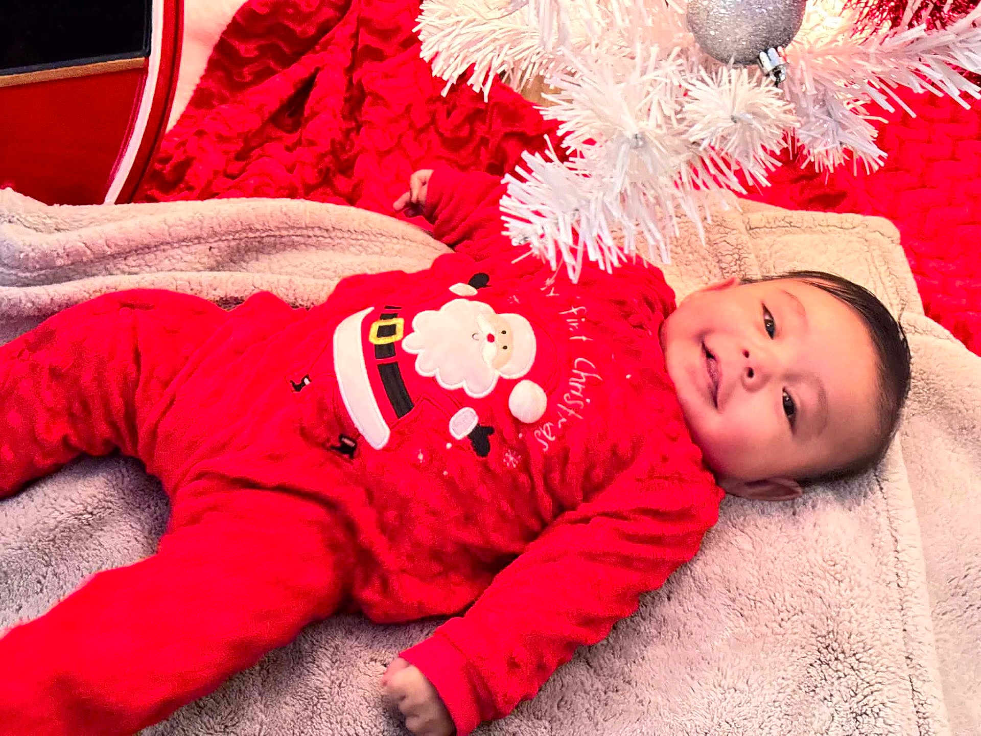 Omar joined the competition — help win amazing prizes! baby, infant, red_clothing, santa_outfit, blanket, christmas_tree, holiday, smile, face, lying_down, cute, festive, soft_texture, indoors, child, happy, christmas_decor, toddler, warm, cozy