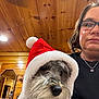 dog, person, glasses, santa_hat, hood, indoor, wood_paneling, portrait, canine, pet, christmas, festive, woman, jewelry, necklace, brown_hair, closeup, casual_clothing, reflection, mirror