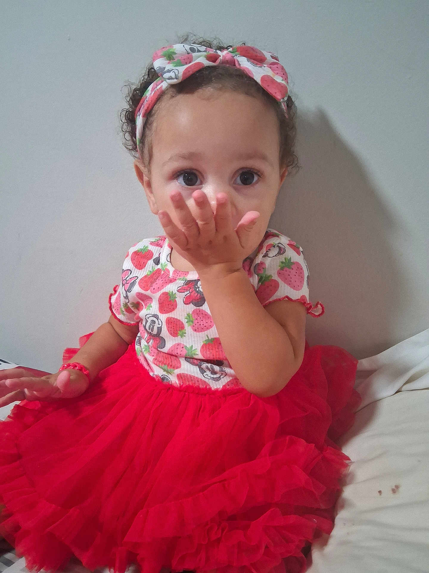 Nakaylia is registered to the contest to win money with this photo: toddler, child, girl, red_tutu, headband, strawberry_pattern, curly_hair, hand_on_face, sitting, innocent_expression, wall, bed, clothing, cute, playful, portrait, indoor, person, young_child, fashion